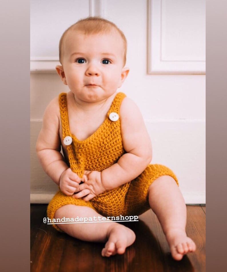 May include: A baby wearing a crocheted yellow romper with white buttons. The romper has a sleeveless design and a wide leg. The baby is sitting on a wooden floor.