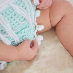 May include: A baby wearing a crocheted outfit with a mint green and white color scheme. The outfit has a fringe detail and white pom-poms.