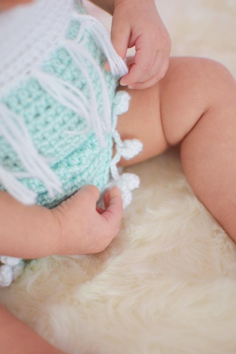 May include: A baby wearing a crocheted outfit with a mint green and white color scheme. The outfit has a fringe detail and white pom-poms.