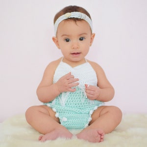 May include: A baby girl wearing a light blue crocheted halter top with white trim and white pom-poms. She is sitting on a white fluffy rug.