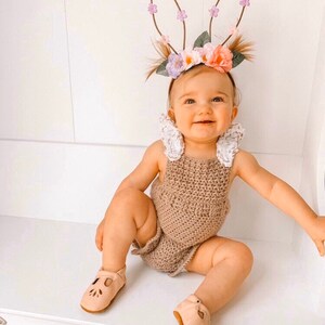 May include: A smiling baby wearing a brown crochet romper, light pink shoes, and a floral bunny ear headband. The headband features brown wire ears adorned with small flowers in shades of pink, purple, and white.