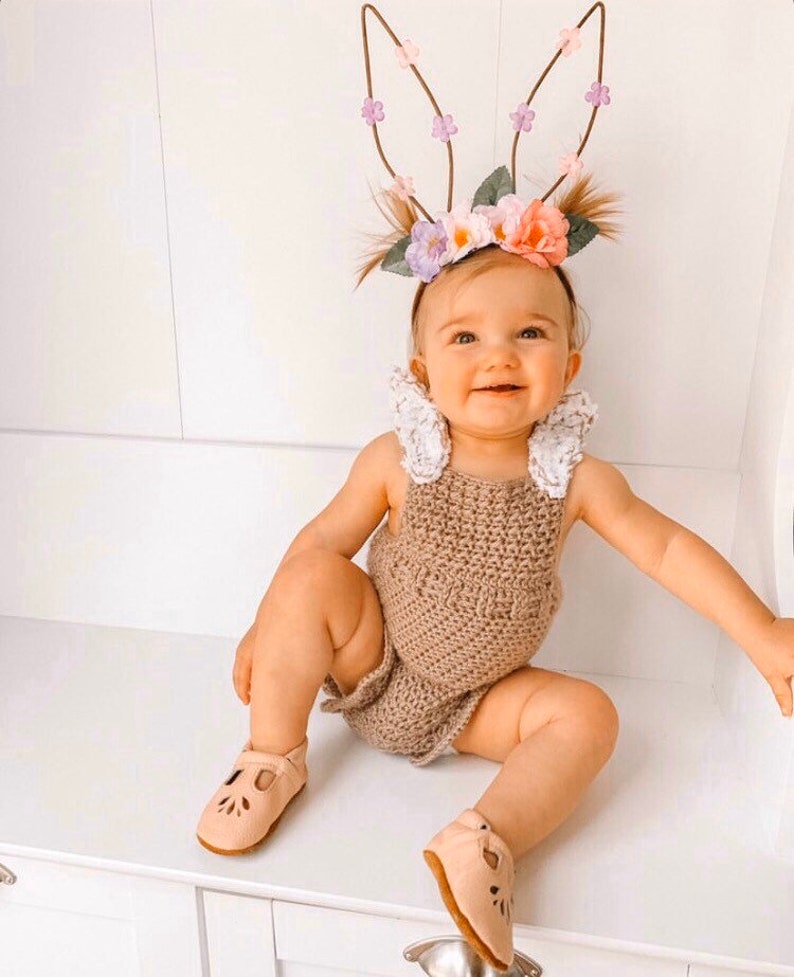 May include: A smiling baby wearing a brown crochet romper, light pink shoes, and a floral bunny ear headband. The headband features brown wire ears adorned with small flowers in shades of pink, purple, and white.
