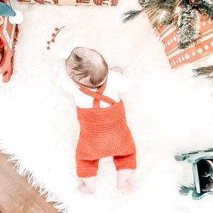 May include: A baby wearing a red crocheted overall with white straps. The baby is lying on a white fluffy rug.