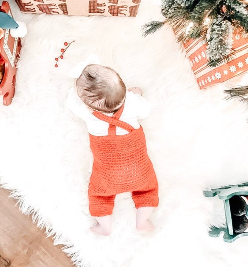 May include: A baby wearing a red crocheted overall with white straps. The baby is lying on a white fluffy rug.