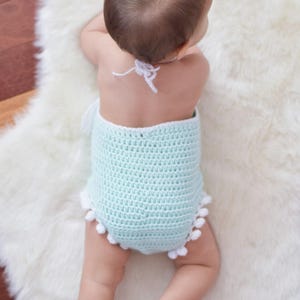 May include: A baby wearing a light blue crocheted romper with white pom-poms around the bottom. The romper has a white crocheted halter strap.