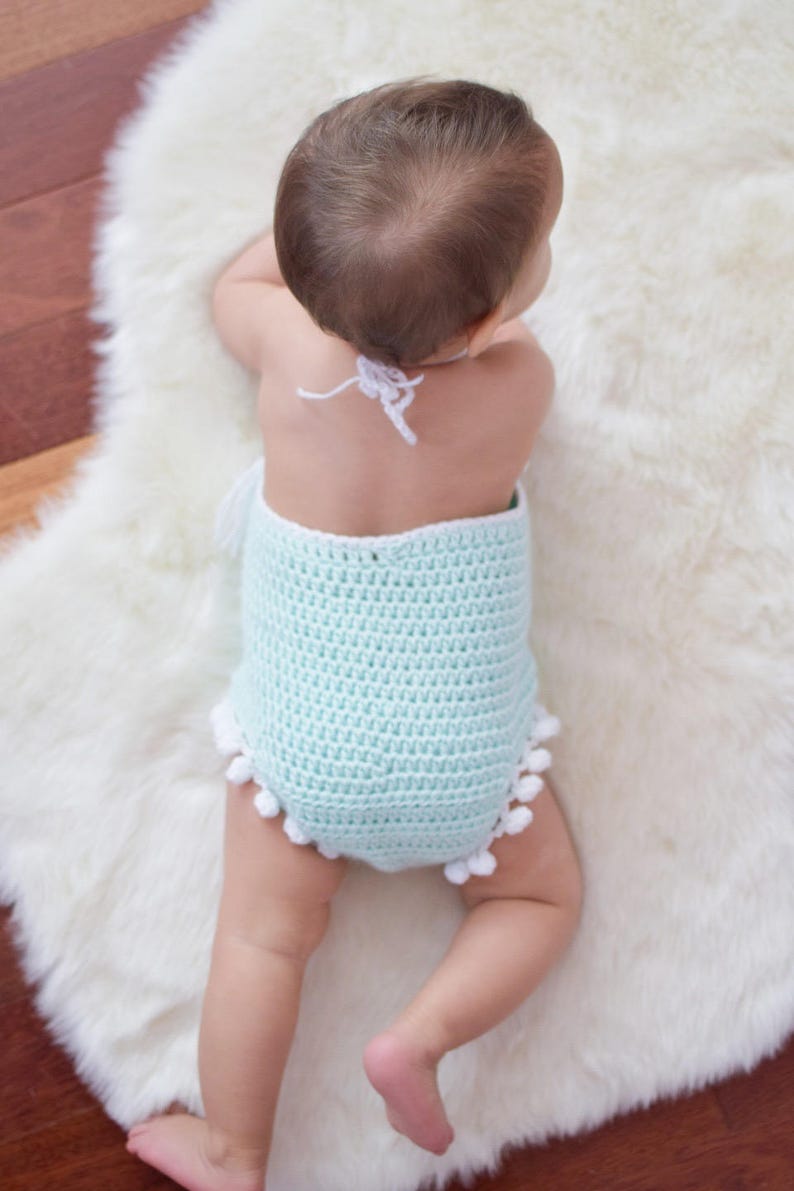 May include: A baby wearing a light blue crocheted romper with white pom-poms around the bottom. The romper has a white crocheted halter strap.