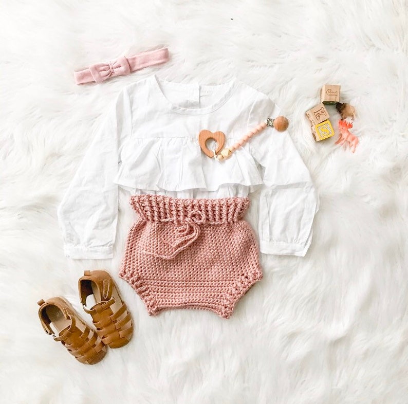 Puede incluir: Unos bloomers de beb&eacute; de crochet rosa con un top blanco con volantes y una diadema rosa. El conjunto est&aacute; colocado sobre una superficie blanca y esponjosa con juguetes de madera.