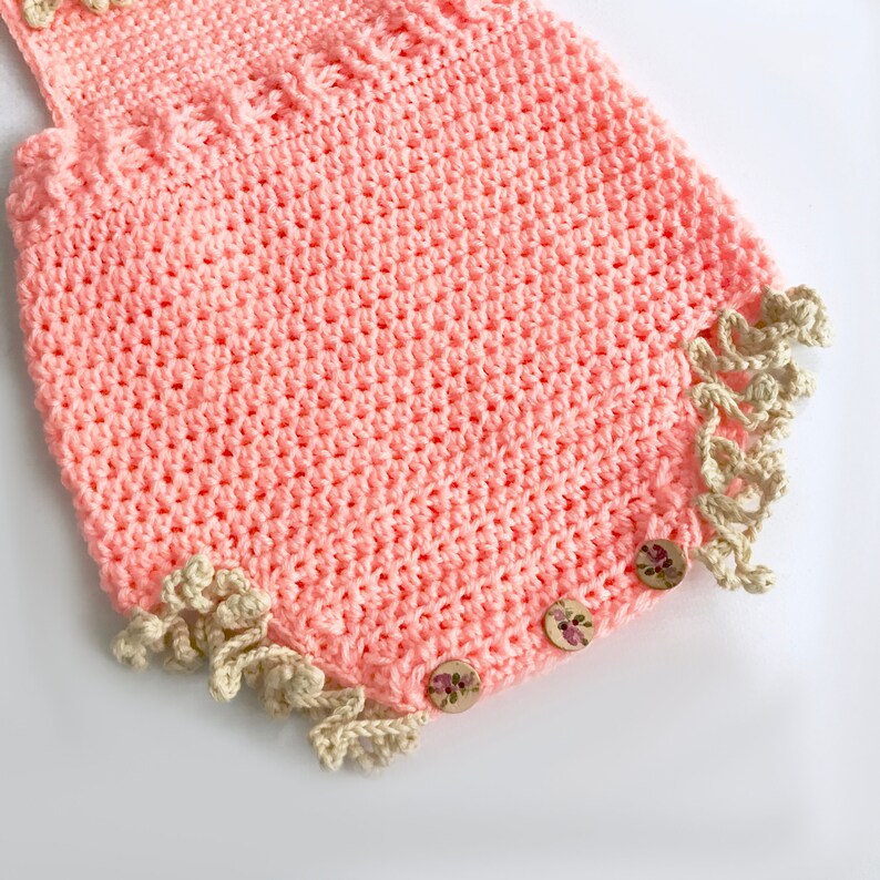 May include: A handmade, peach-colored crochet romper with a textured stitch pattern. The edges are trimmed with a cream-colored, ruffled crochet detail. Wooden buttons with floral designs secure the garment, showcasing a delicate, handcrafted aesthetic.