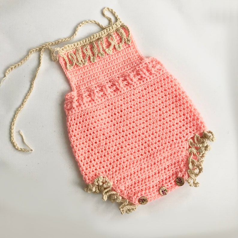 May include: A handmade, light pink crochet romper with beige trim and straps. The romper features decorative edging, wooden buttons, and a textured stitch pattern. The straps are braided, and the romper is designed for a baby or toddler.