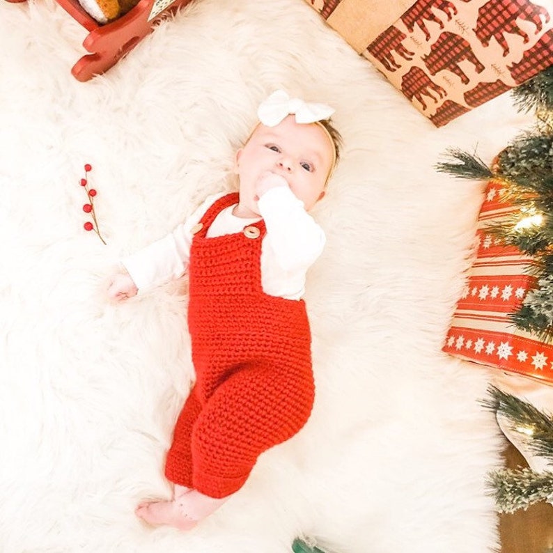 May include: A baby wearing a red crocheted overall with a white bow headband. The baby is lying on a white fluffy surface.