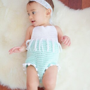 May include: A baby wearing a crocheted white and light blue romper with white fringe and white pom-poms. The baby is wearing a white headband with a lace design.