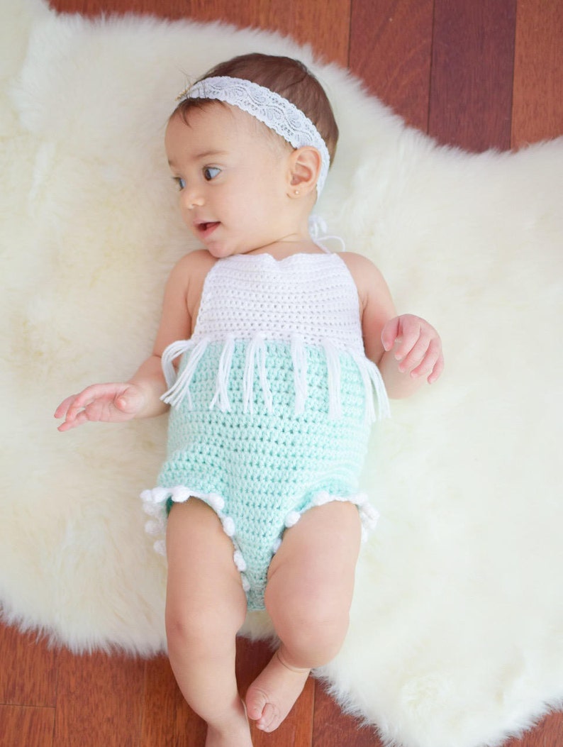 May include: A baby wearing a crocheted white and light blue romper with white fringe and white pom-poms. The baby is wearing a white headband with a lace design.