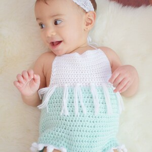 May include: A baby wearing a light green crocheted romper with white crocheted trim and white pom-poms. The romper has a white crocheted halter top with fringe.