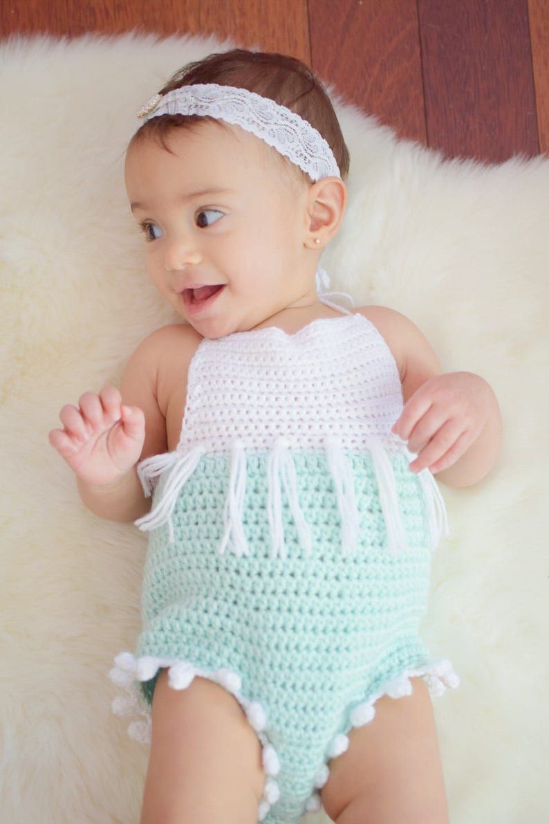 May include: A baby wearing a light green crocheted romper with white crocheted trim and white pom-poms. The romper has a white crocheted halter top with fringe.
