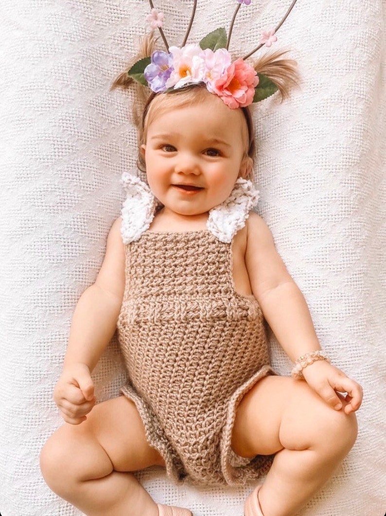 May include: A smiling baby wearing a handmade, light brown crochet romper with white lace accents. The baby is also wearing a floral headband with pink, purple, and white flowers and a beaded bracelet. The baby is lying on a white textured blanket.