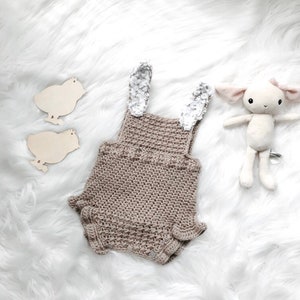 May include: A crocheted baby romper in a neutral tan color, featuring bunny ears with a floral pattern. The romper is accompanied by a plush lamb toy and two wooden chick cutouts, all set against a white, fluffy background.