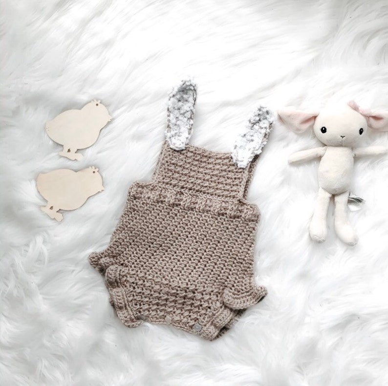 May include: A crocheted baby romper in a neutral tan color, featuring bunny ears with a floral pattern. The romper is accompanied by a plush lamb toy and two wooden chick cutouts, all set against a white, fluffy background.