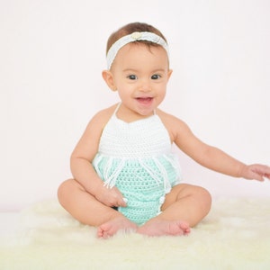 May include: A baby girl wearing a white and light green crocheted dress with a white headband. The baby is sitting on a white fluffy rug.