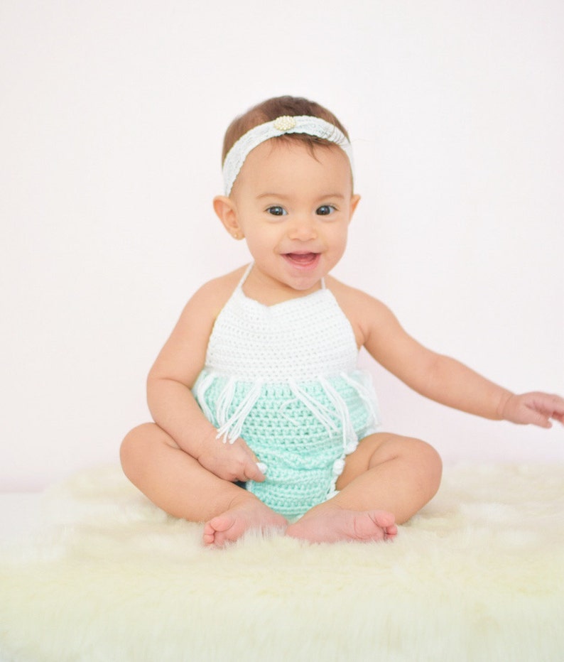 May include: A baby girl wearing a white and light green crocheted dress with a white headband. The baby is sitting on a white fluffy rug.