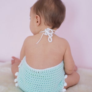 May include: A baby wearing a light blue crocheted romper with white trim and pom-poms. The romper has a white crocheted halter strap.