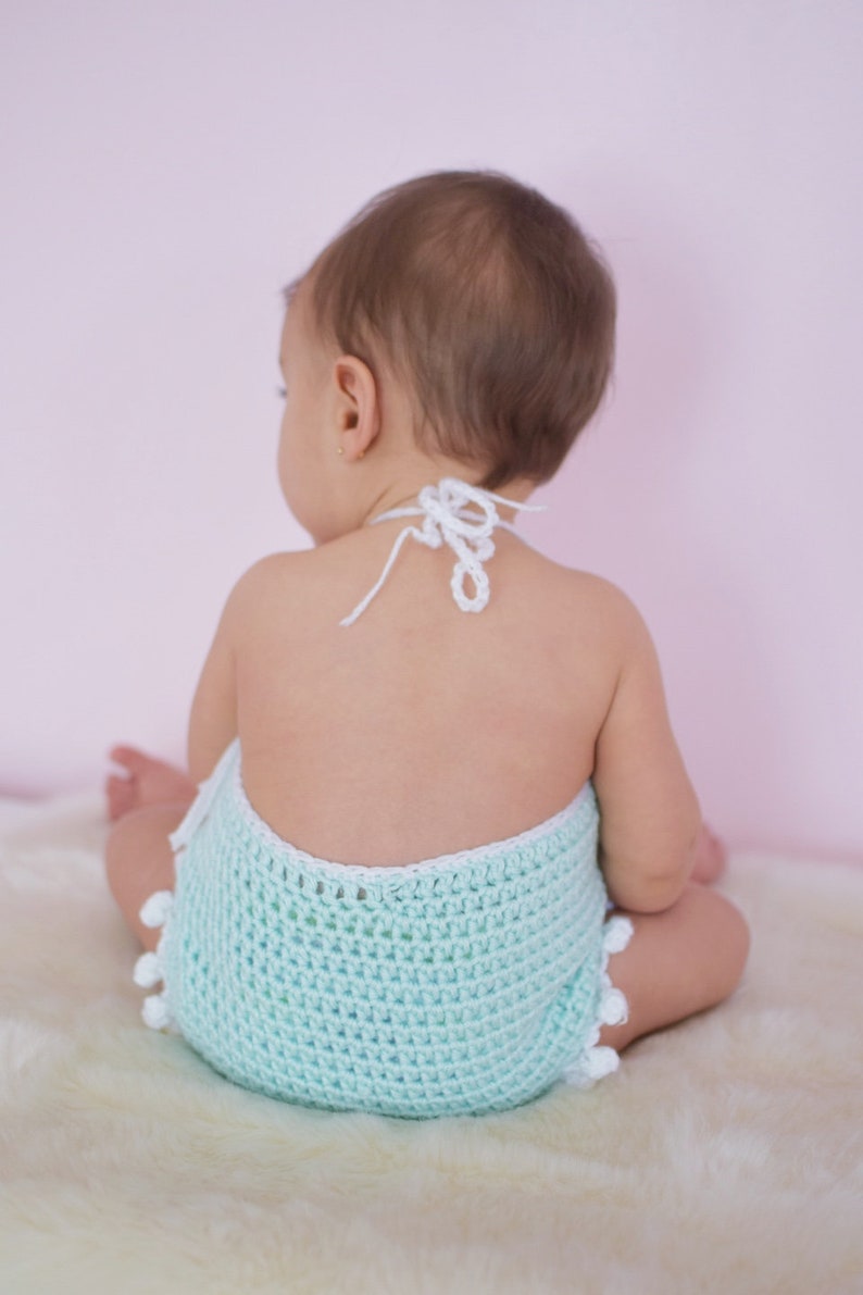 May include: A baby wearing a light blue crocheted romper with white trim and pom-poms. The romper has a white crocheted halter strap.