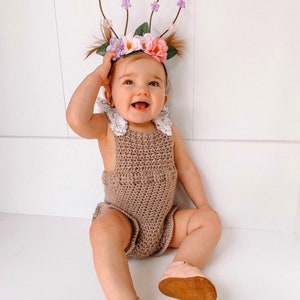 May include: A smiling baby wearing a brown crochet romper with white lace trim and pink leather shoes. The baby is also wearing a floral headband with bunny ears, perfect for Easter or a spring photoshoot.