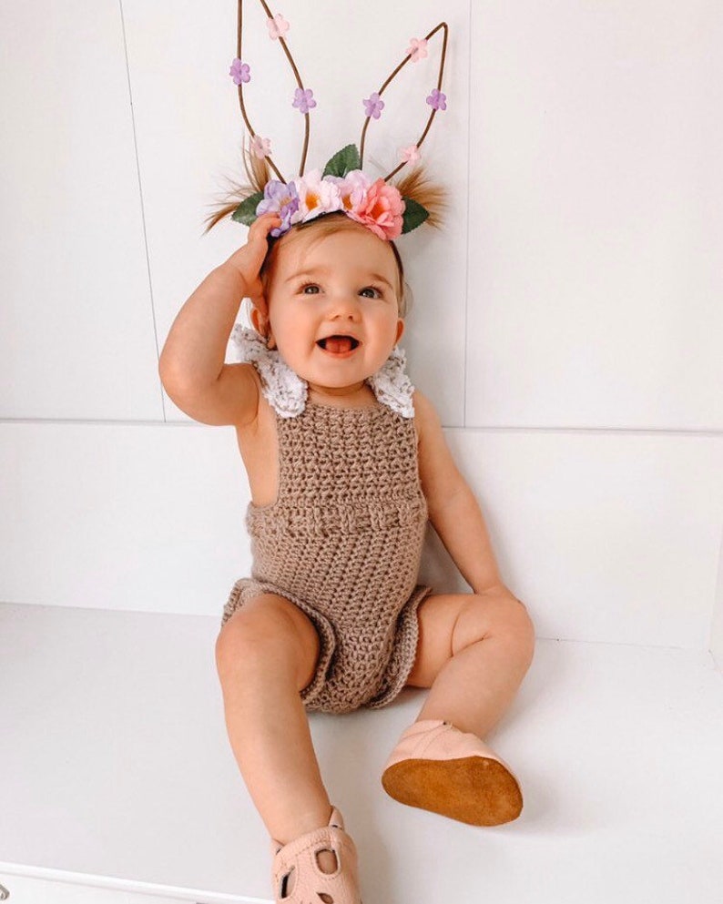 May include: A smiling baby wearing a brown crochet romper with white lace trim and pink leather shoes. The baby is also wearing a floral headband with bunny ears, perfect for Easter or a spring photoshoot.