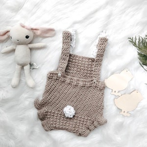 May include: A crocheted baby romper in a neutral tan color with white lace accents and a fluffy white tail. A stuffed bunny and wooden chick cutouts are also in the image, suggesting a baby or Easter theme.