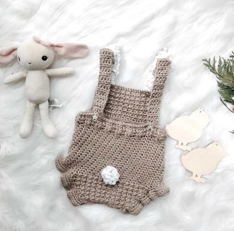 May include: A crocheted baby romper in a neutral tan color with white lace accents and a fluffy white tail. A stuffed bunny and wooden chick cutouts are also in the image, suggesting a baby or Easter theme.