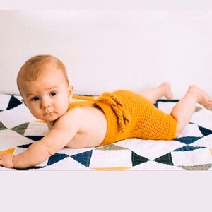 May include: A baby wearing a yellow crocheted romper with a bow on the back, lying on a white and black patterned quilt.