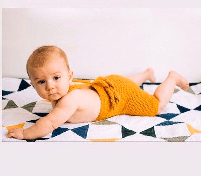 May include: A baby wearing a yellow crocheted romper with a bow on the back, lying on a white and black patterned quilt.