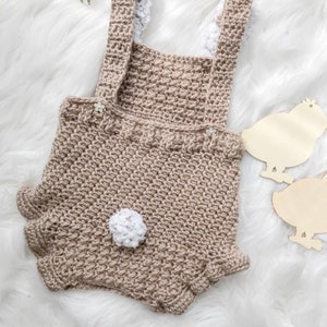 May include: A handmade crochet romper in a neutral beige color, featuring a textured stitch pattern. The romper has suspender straps with white trim, small flower accents, and a white pom-pom detail. The design includes ruffled edges on the shorts.