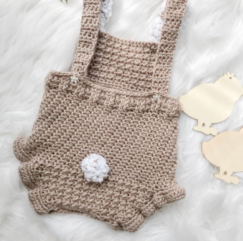 May include: A handmade crochet romper in a neutral beige color, featuring a textured stitch pattern. The romper has suspender straps with white trim, small flower accents, and a white pom-pom detail. The design includes ruffled edges on the shorts.