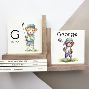 May include: A set of two wooden blocks. One block features the letter G and a cartoon leopard wearing a blue shirt and cap holding a golf club. The other block has the name George and a cartoon monkey wearing a blue shirt and cap holding a golf club.