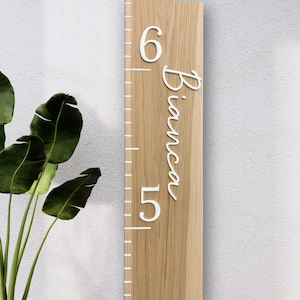 May include: A wooden growth chart with white numbers and the name "Bianca" in cursive. The chart has a measuring scale and is made of light-colored wood. A green plant is visible in the background.