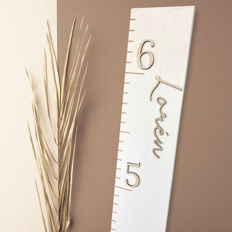 May include: White wooden growth chart with a ruler and the name "Lauren" written in cursive. The numbers "6" and "15" are also visible on the chart.