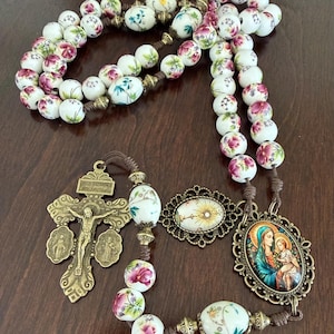 May include: A rosary featuring white ceramic beads decorated with pink floral patterns and green foliage, separated by small bronze-toned metal spacers. It includes a bronze-toned crucifix with the inscription JESUS NAZARENUS REX JUDAEORUM and an oval pendant depicting a mother and child.