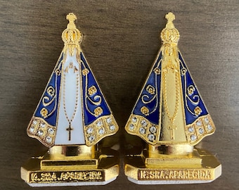 Puede incluir: Dos figuritas doradas de Nuestra Señora de Aparecida, un icono católico. Las figuritas llevan túnicas azules y blancas con ribetes dorados y una corona. La base de cada figurita es dorada y tiene las palabras "Nossa Senhora Aparecida" escritas en ella.