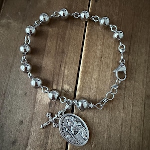 May include: A silver-toned bracelet featuring a series of round beads and a decorative pendant. The pendant depicts a religious figure and a small cross charm. The bracelet is secured with a clasp.