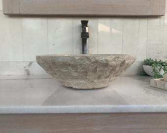 May include: A large, round, light brown stone sink with a rough, textured surface. The sink is set on a white countertop with a single faucet.