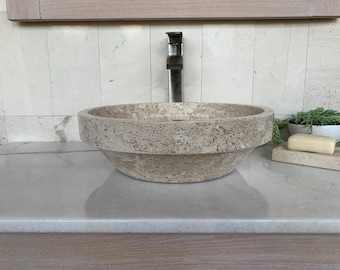 May include: A round, beige, natural stone vessel sink with a textured, porous surface. The sink features a wide, flat rim and a tapered base, positioned beneath a tall, metallic faucet. The basin is crafted from a single piece of stone.