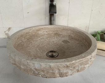 May include: A round vessel sink crafted from beige natural stone with a textured, chiseled exterior and a smooth interior basin. The sink features a central metallic drain plug and is positioned beneath a tall, dark metal faucet.