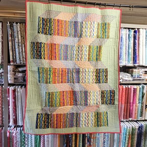 May include: A handmade quilt with a light green background and a red border. The quilt features a geometric design of colorful vertical stripes in various colors, including yellow, blue, and red. The quilt is displayed in a fabric store.