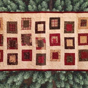 May include: A beige table runner with a red border and a patchwork design of squares in shades of brown, red, and green.