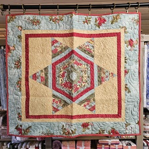 May include: A square quilt with a floral pattern in shades of blue, red, and beige. The quilt features a central diamond design and is bordered by a floral print. The quilt is hanging from a black rod.