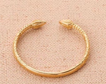 May include: A gold-colored open cuff bracelet featuring a textured, patterned surface along the band and two pointed, tapered ends. The metal has a polished finish, and the design is circular with a gap at the top for an adjustable fit.