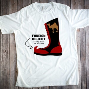 May include: White t-shirt with a red and black boot graphic. The boot has a camel on it and the text "FOREIGN OBJECT" with the words "Stomp boot to activate" underneath.