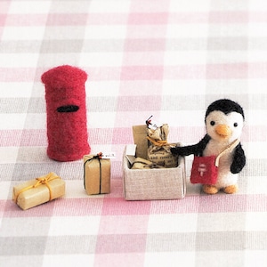 Op de afbeelding: Een klein pinguïnfiguurtje dat een rode tas vasthoudt met een wit Japans karakter erop. De pinguïn staat naast een klein doosje gevuld met bruine papieren pakketten. Een rode brievenbus staat op de achtergrond.