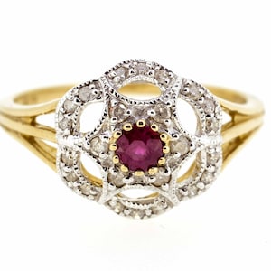 May include: A gold ring with a red ruby gemstone surrounded by a halo of smaller diamonds. The ring has a delicate, intricate design.
