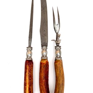 Può includere: Un set di utensili da intaglio antichi, tra cui un coltello, una forchetta e un'acciarino. Le lame sono argentate e i manici sono in materiale marrone-rossastro con dettagli argentati. Il set è presentato su sfondo bianco.